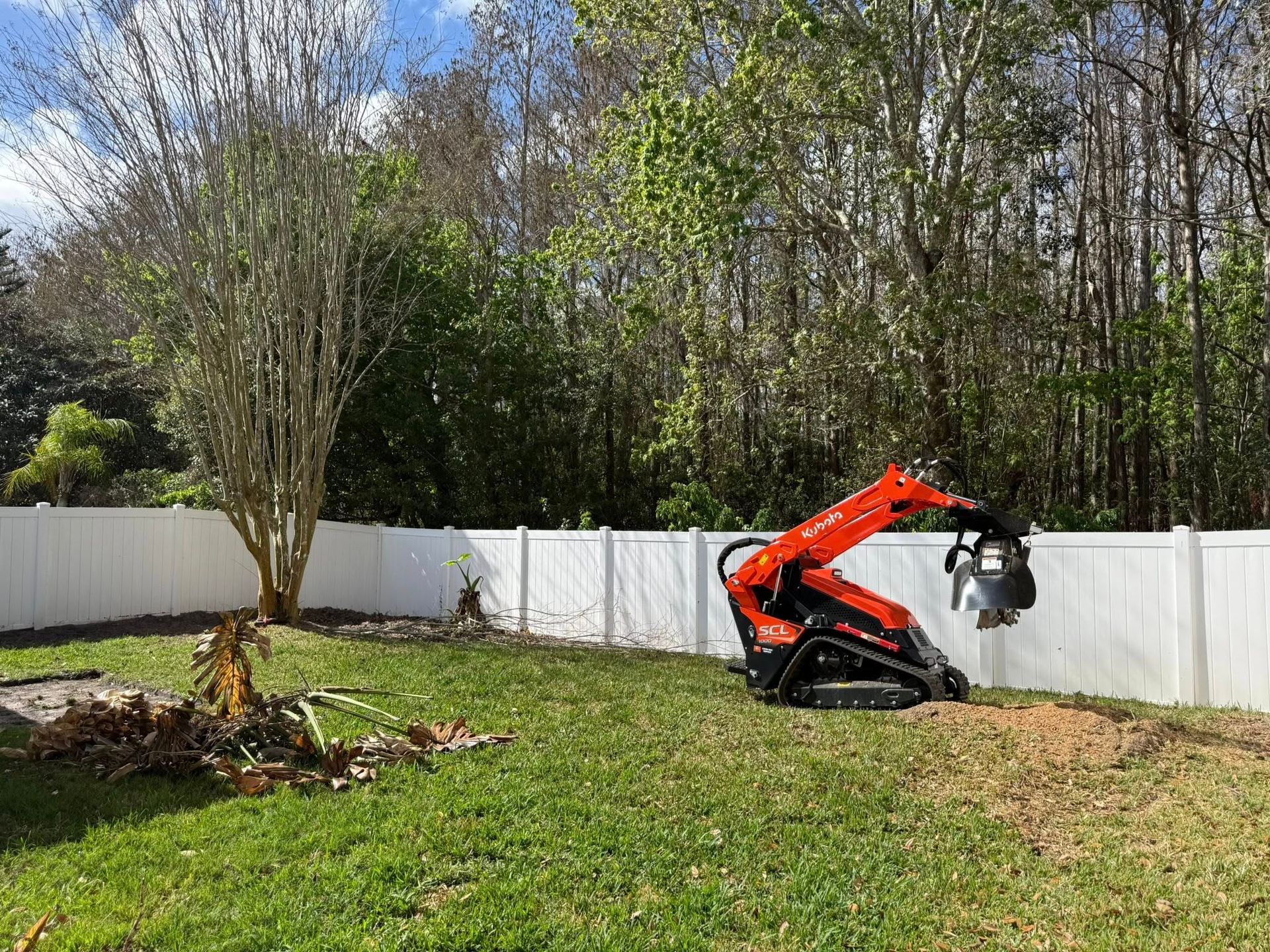An orange tracked skid steer loader operates in a grassy backyard near a white fence and a row of trees.