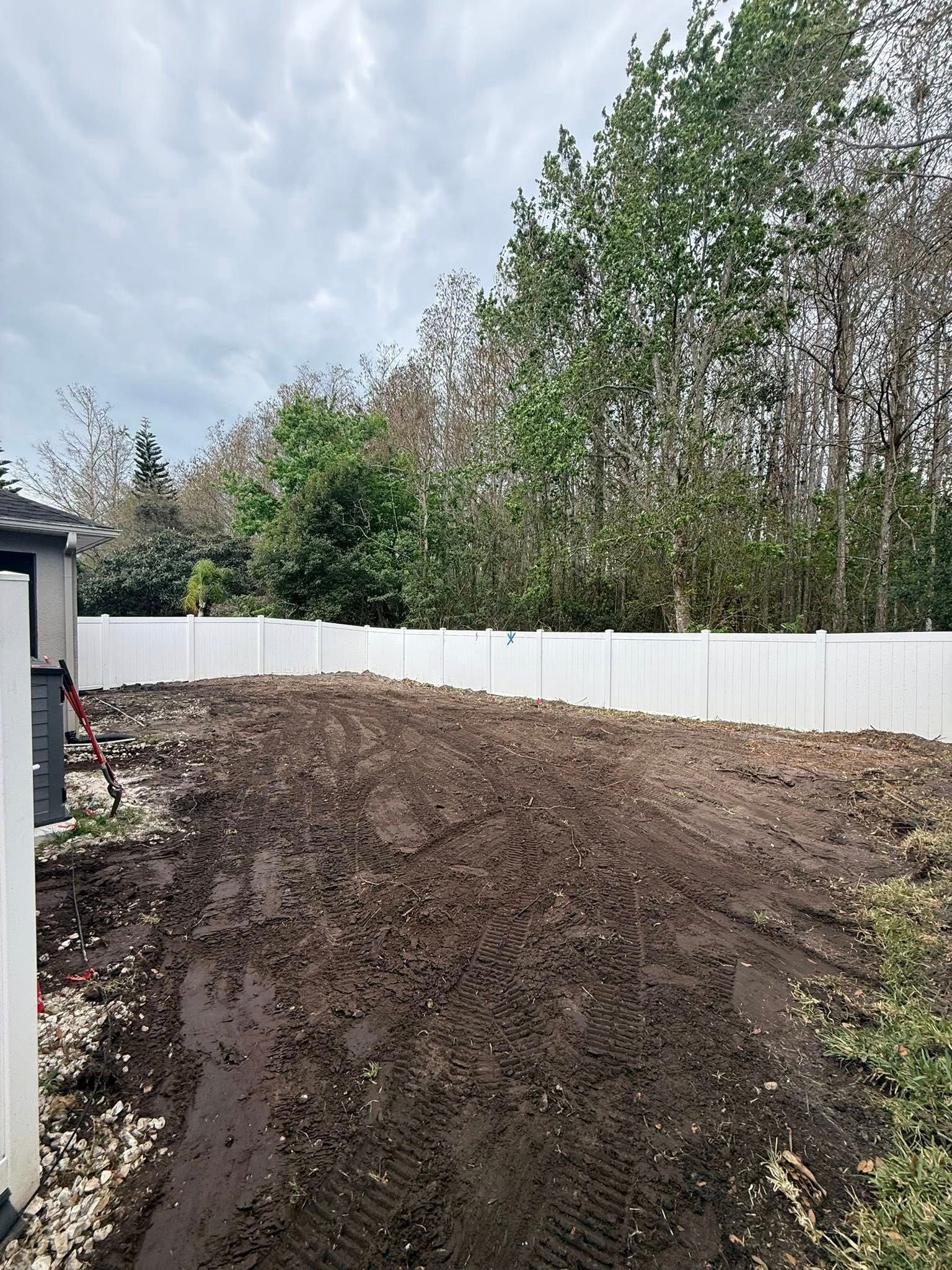 A freshly cleared, muddy backyard area enclosed by a white vinyl fence with a line of trees in the background.