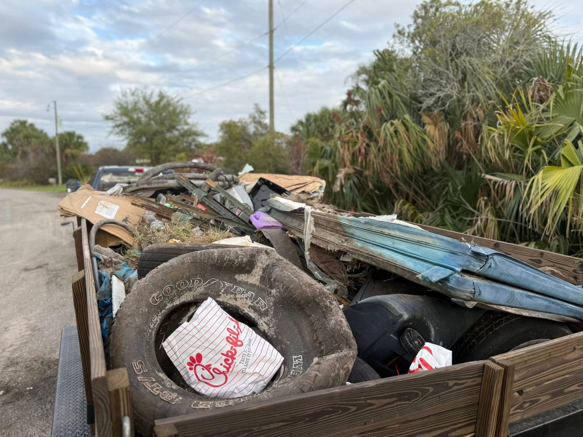 A trailer bed filled with various trash, including old tires, cardboard, and blue metal siding, parked on a roadside.