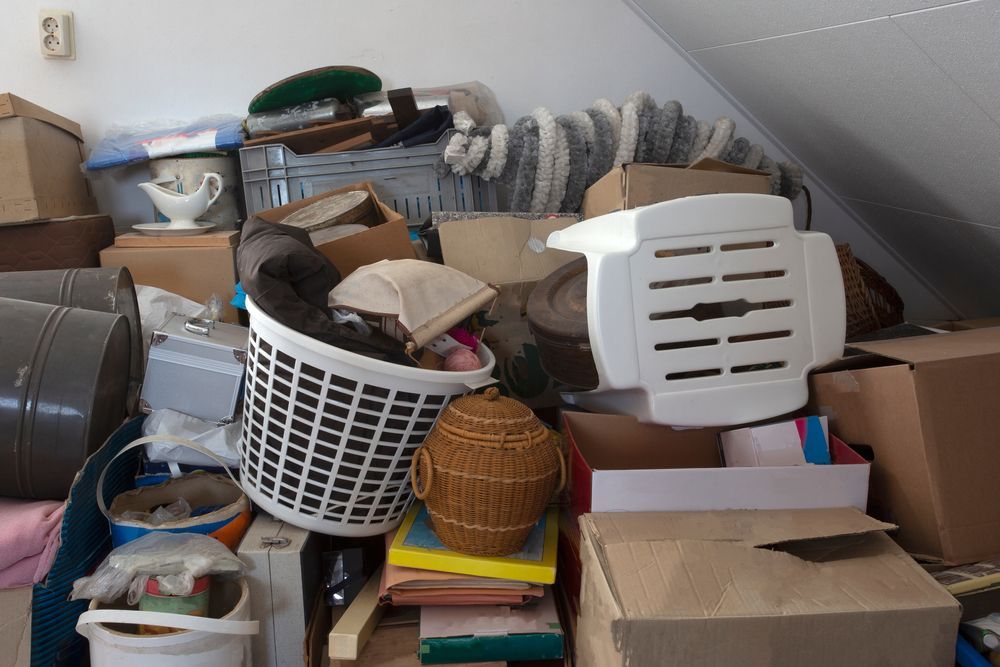 A cluttered attic filled with stacked cardboard boxes, a white plastic laundry basket, a woven basket, and misc items.