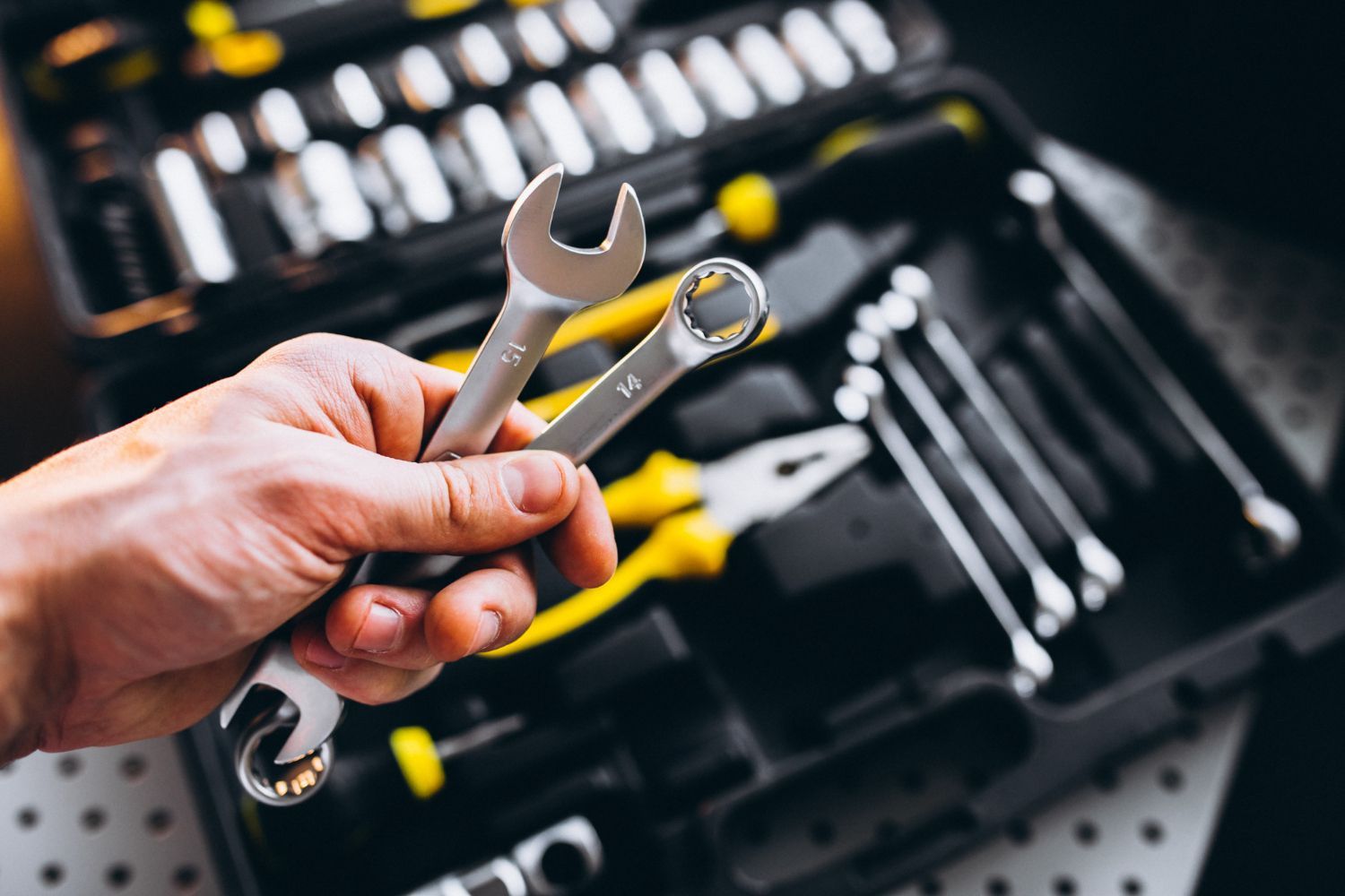 Hand holding two wrenches in front of a partially open toolbox filled with tools.
