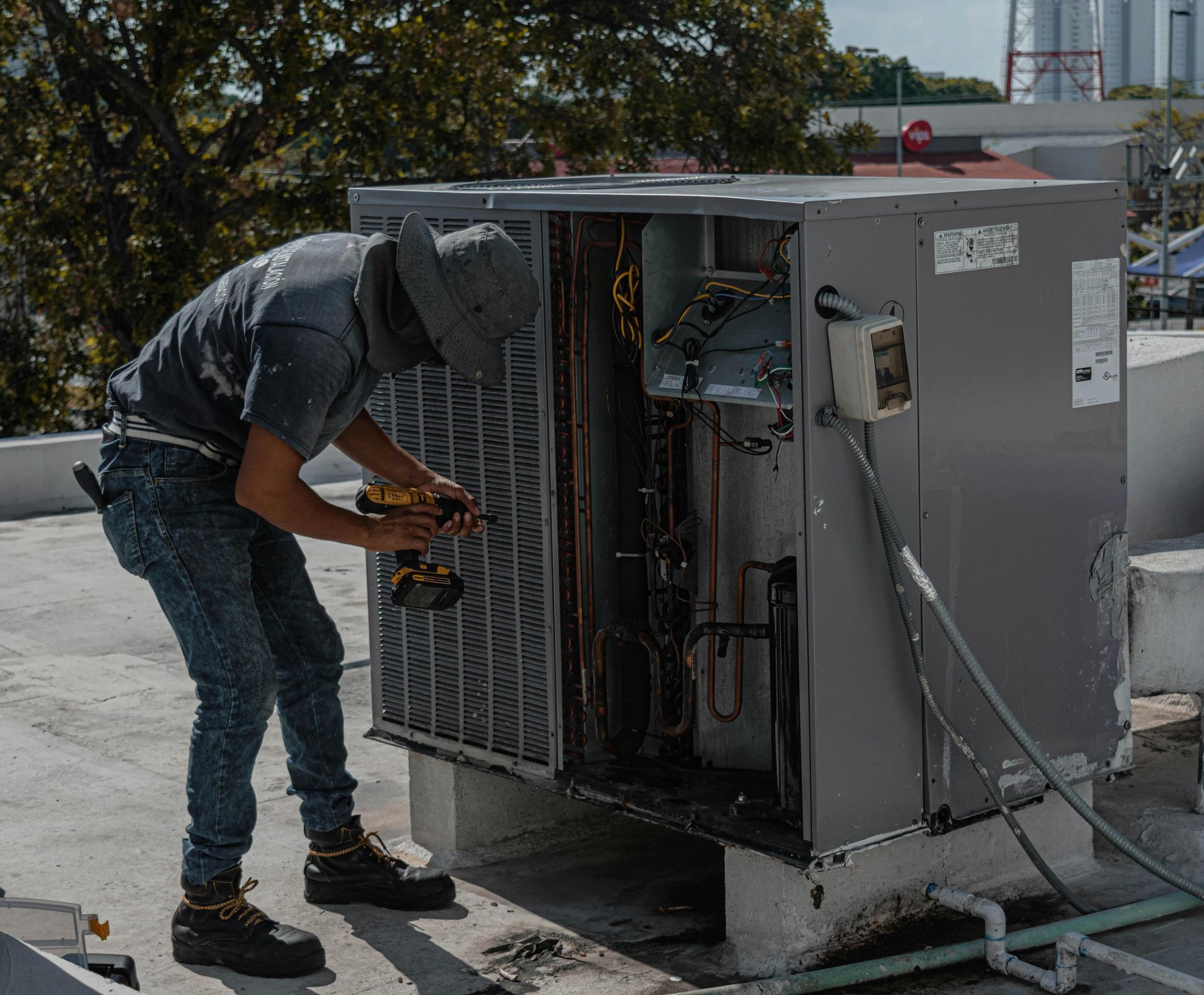 HVAC technician on a rooftop working on an air conditioning unit using a drill.