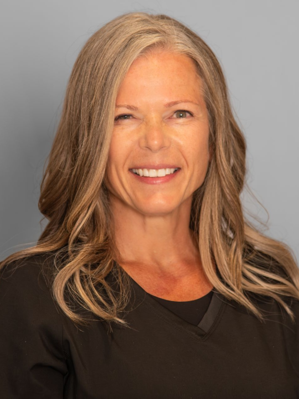 Smiling woman with long blonde hair, wearing a black top, against a gray background.