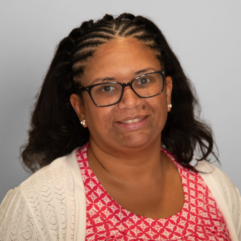 Woman with glasses smiles at the camera; she has dark braids and wears a white sweater over a patterned pink top.