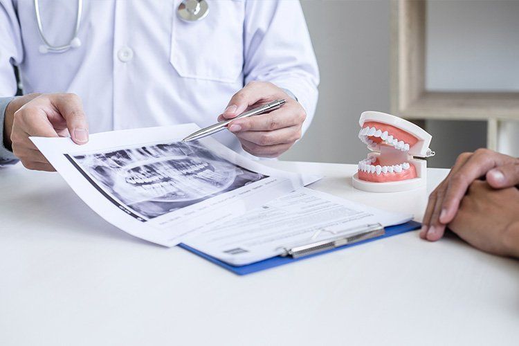 Dentist pointing to a dental X-ray, explaining it to a patient, next to a dental model, in an office.