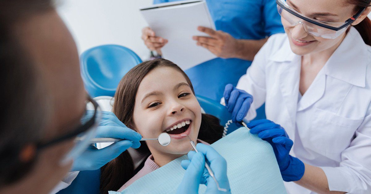 Girl at dentist smiling during a checkup. Dentist and assistant in scrubs, using tools, blue gloves, and glasses.