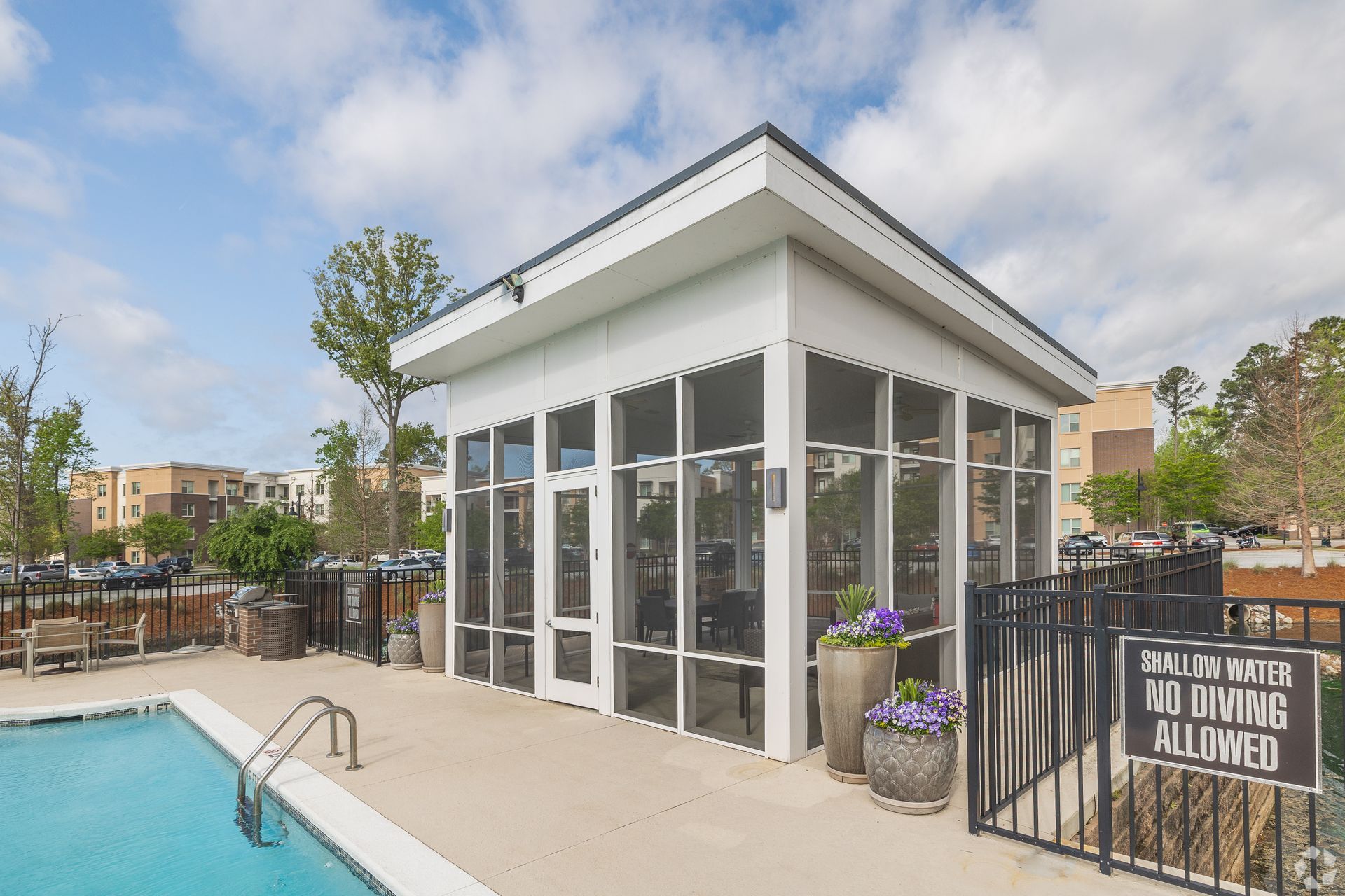 Poolside cabana with glass walls and white trim beside a swimming pool, with a fenced patio and potted flowers.