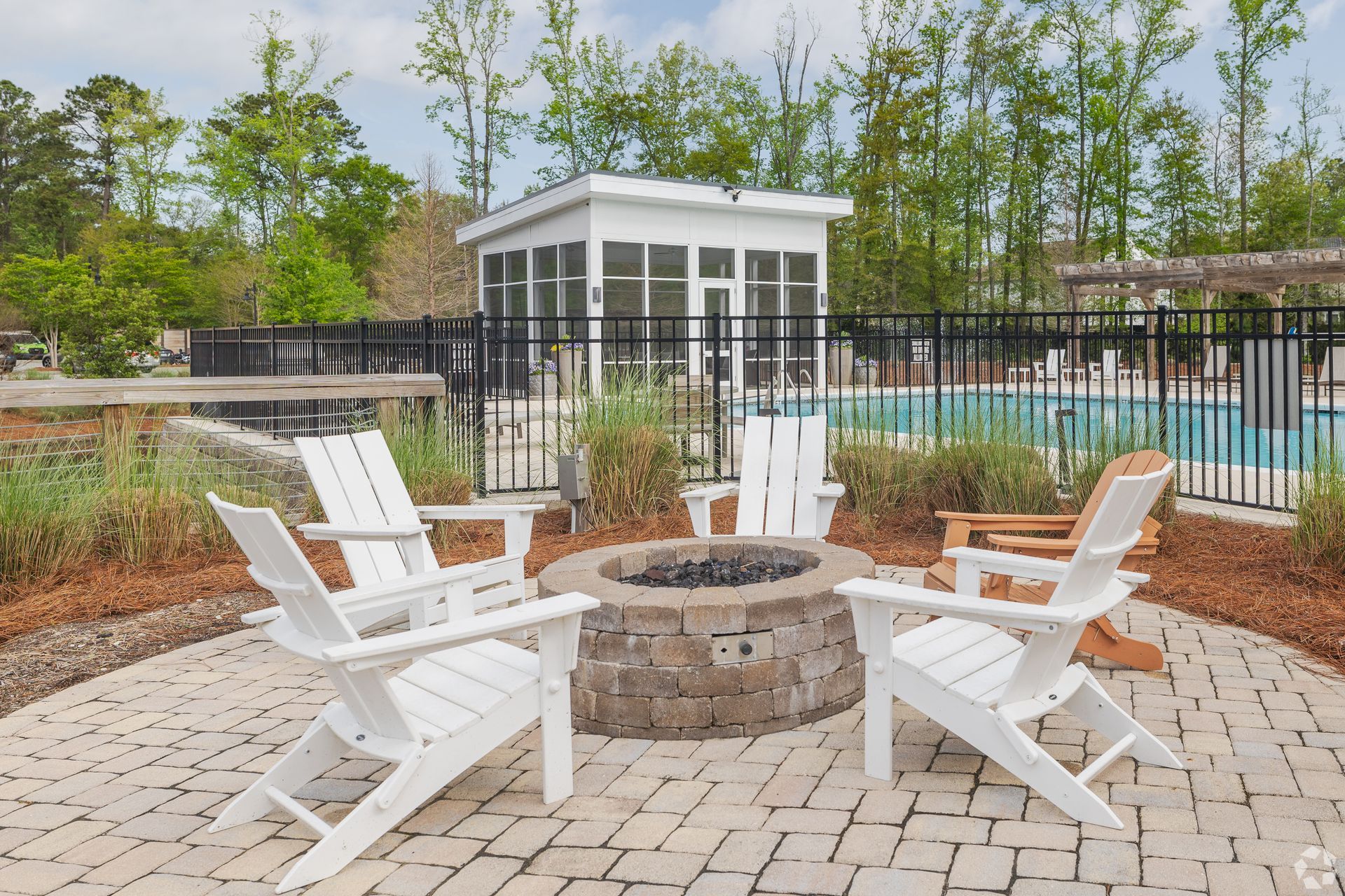 White Adirondack chairs around a stone fire pit beside a fenced pool and small pavilion outdoors.