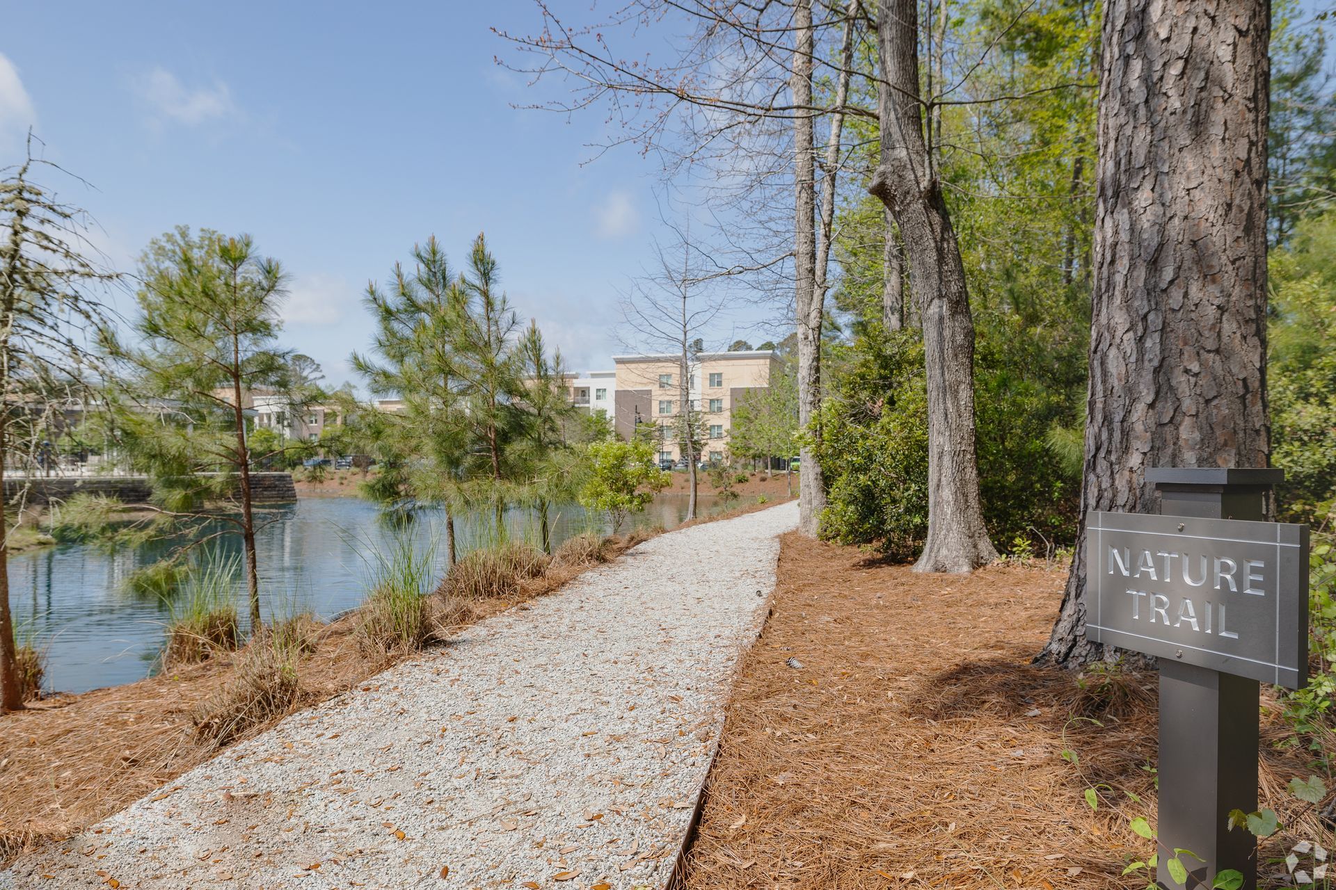 Wooded nature trail beside a pond, with apartment buildings in the distance and a trail sign in front