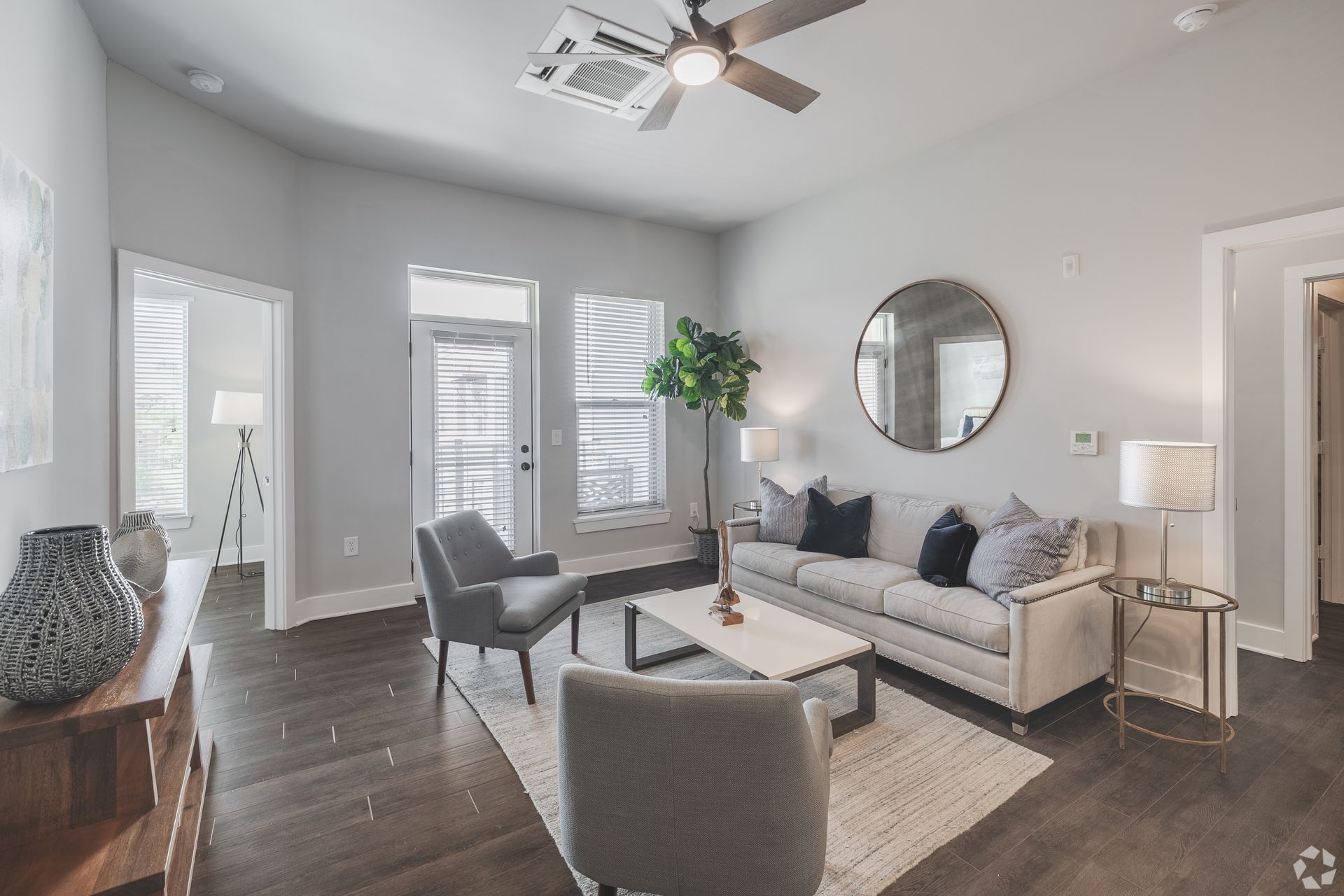 Bright living room with gray sofa, two chairs, round mirror, large windows, and dark wood floors