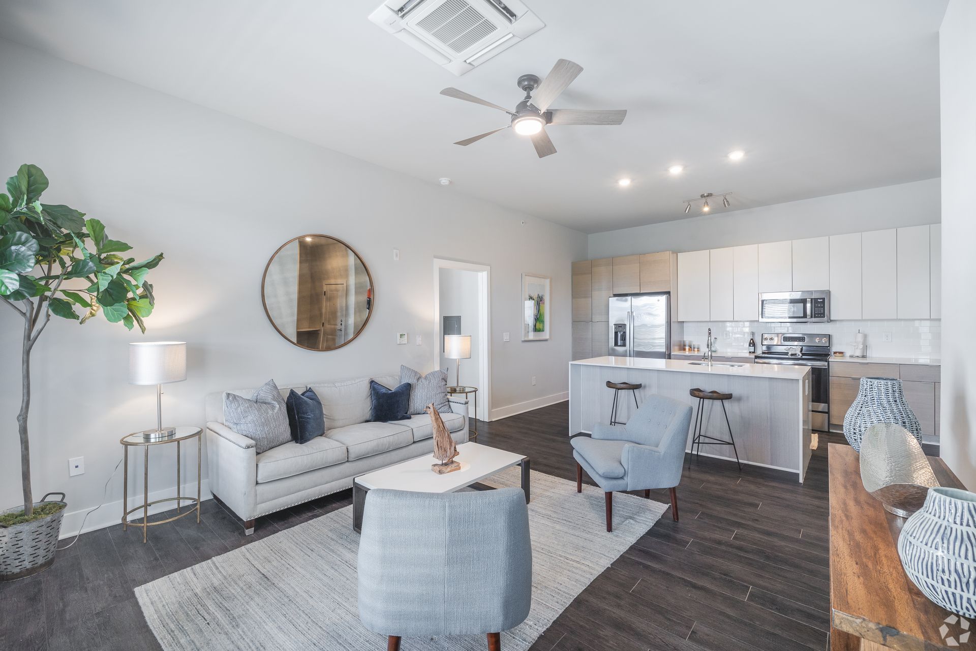 Bright open-concept living room and kitchen with gray sofa, round mirror, and white cabinetry