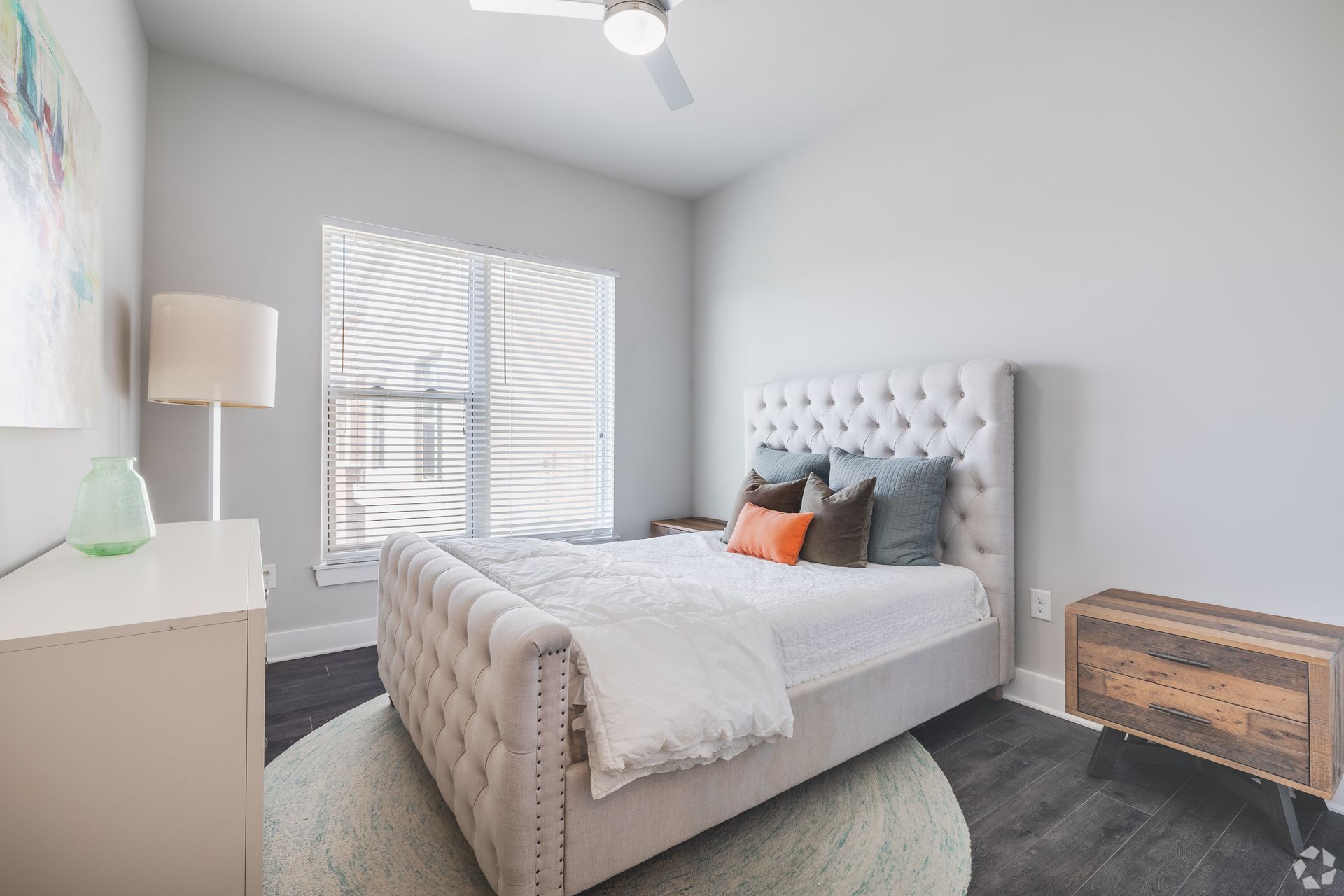Bright bedroom with white tufted bed, window blinds, floor lamp, and wicker chest on a dark rug.