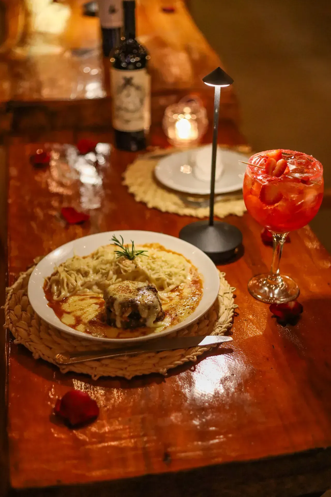 Uma mesa rústica de restaurante com um prato de massa, um coquetel vermelho e uma garrafa iluminada por um pequeno abajur.