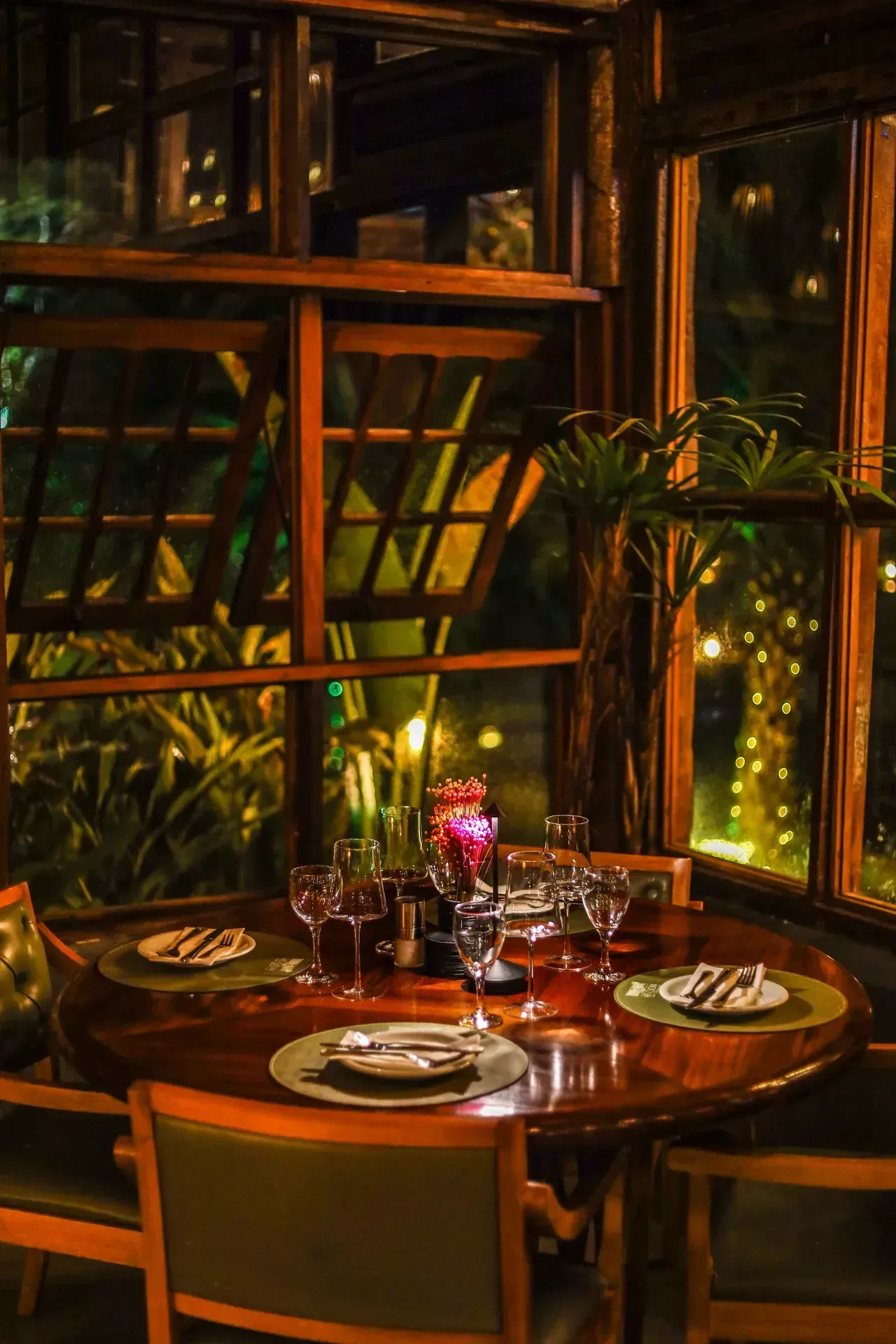 Mesa aconchegante à luz de velas em um restaurante perto da janela, com pratos e copos posta em meio a uma vegetação exuberante.