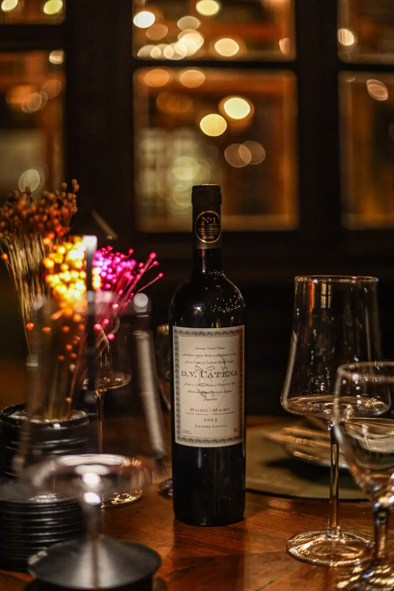 Garrafa e taça de vinho sobre uma mesa de restaurante à luz de velas, com luzes bokeh quentes ao fundo.