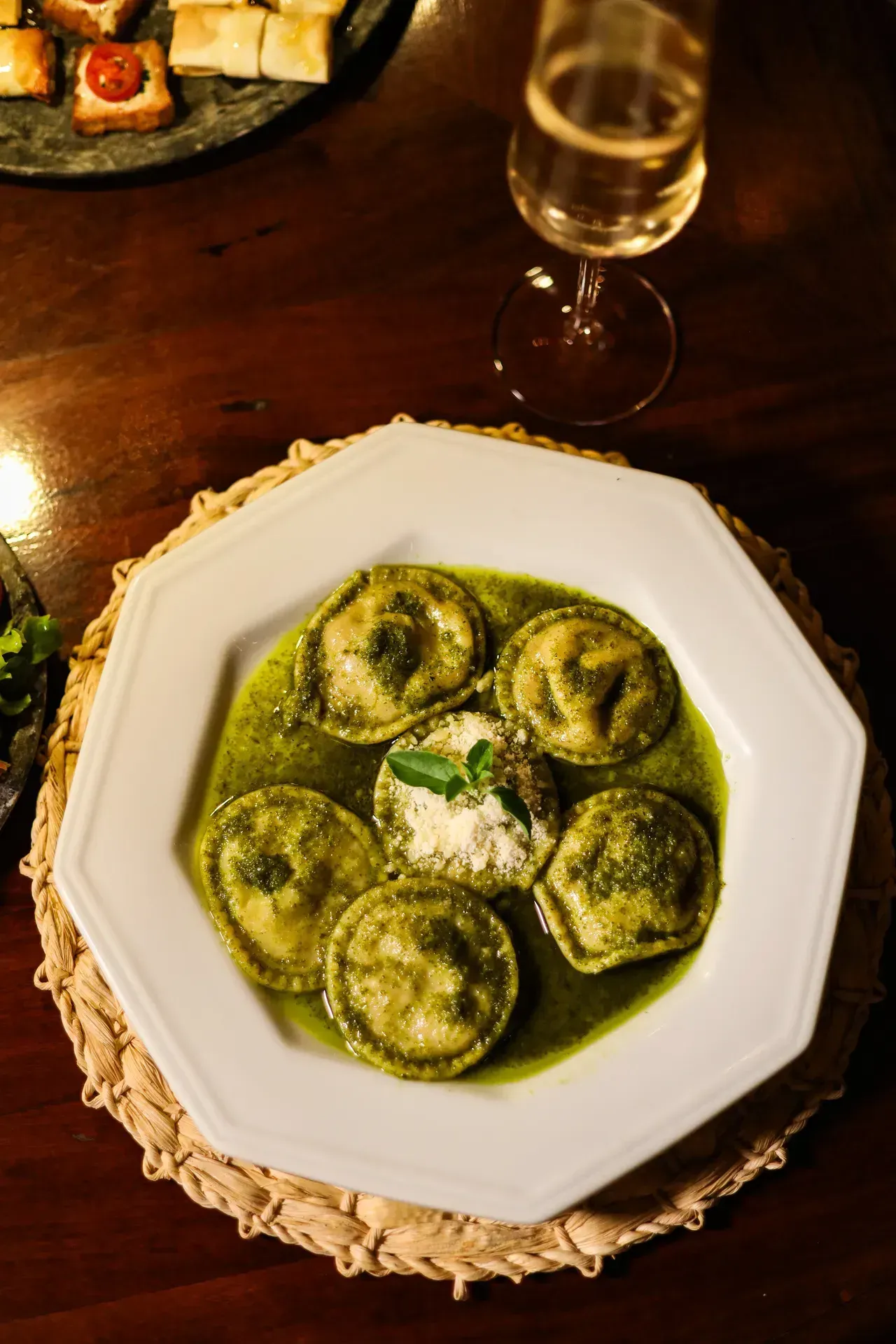 Prato de ravióli verde ao molho pesto sobre a mesa, acompanhado de uma taça de vinho.