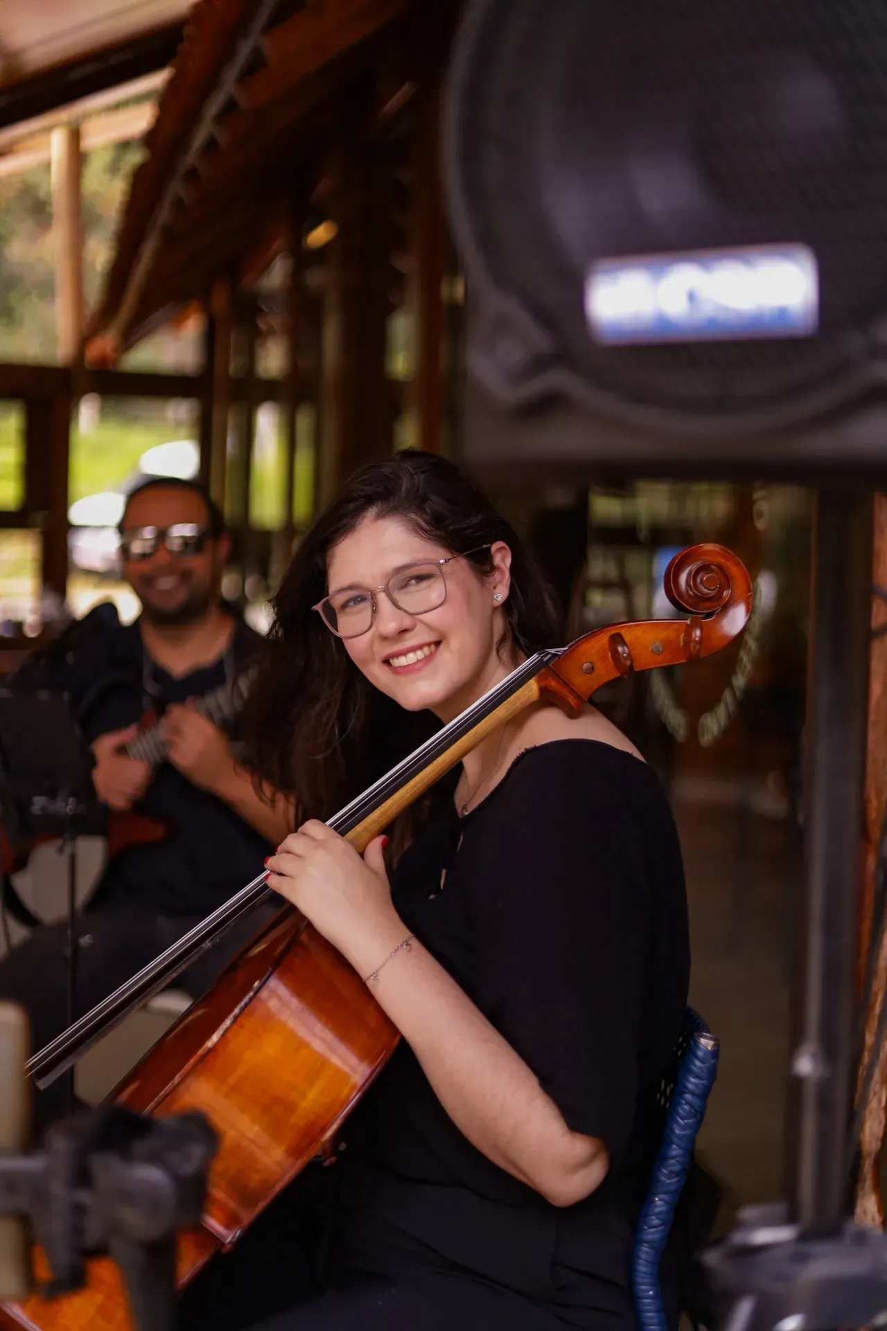 Mulher sorrindo enquanto toca violoncelo em um local ao ar livre, com um músico desfocado ao fundo.