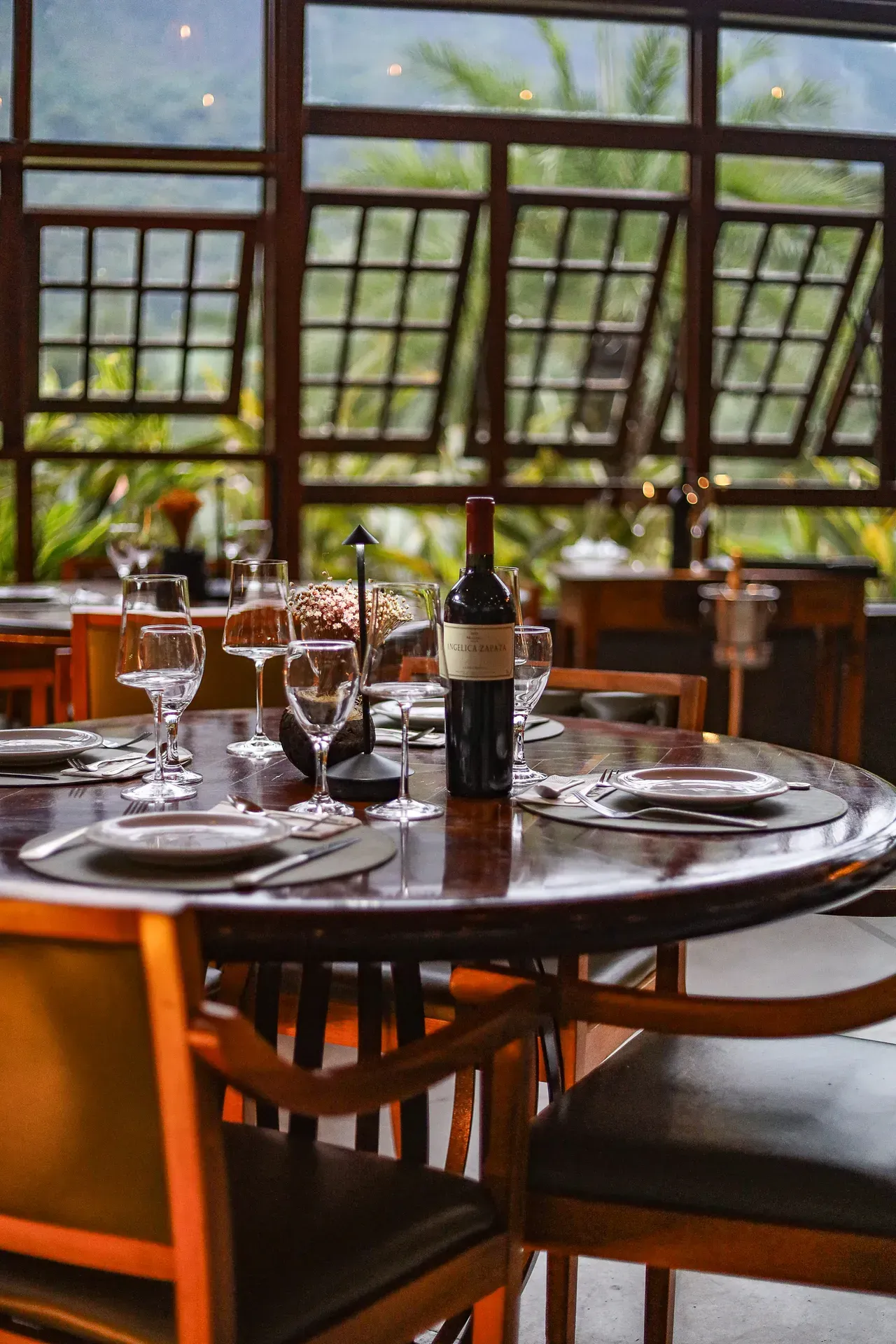 Mesa posta em um restaurante ensolarado, com taças e uma garrafa de vinho junto a grandes janelas.