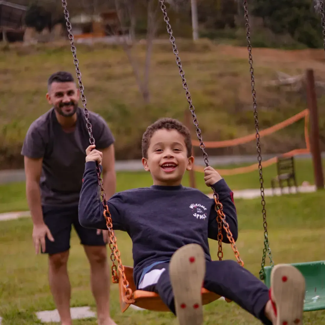 Criança sorrindo em um balanço enquanto um adulto está atrás, em um parque.