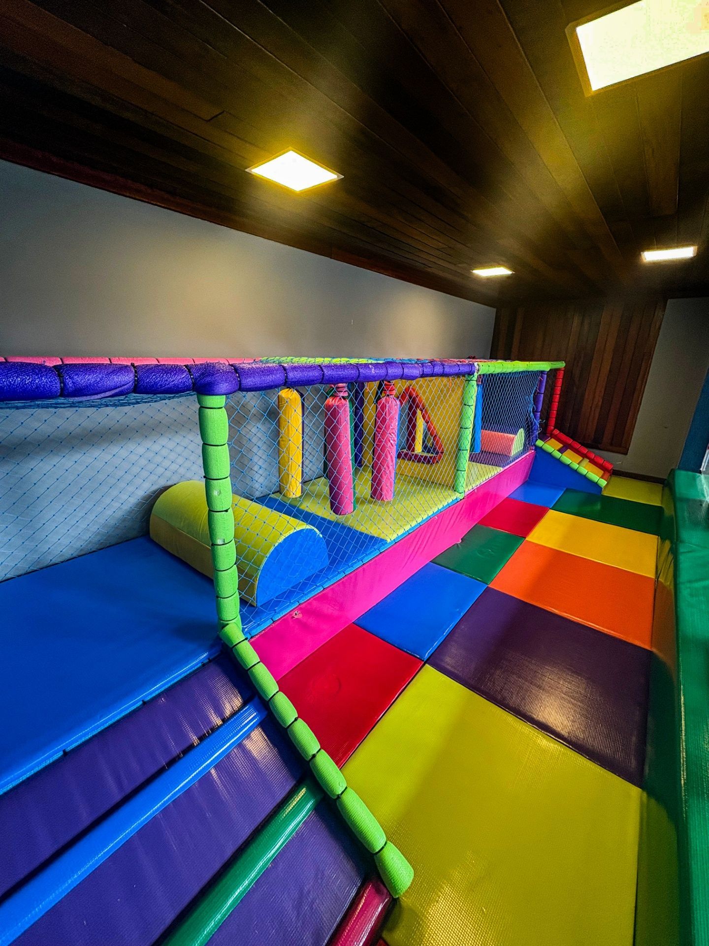 Túnel infantil colorido para brincar em ambientes internos, com rampas acolchoadas, tapetes e obstáculos suspensos sob luzes brilhantes no teto.