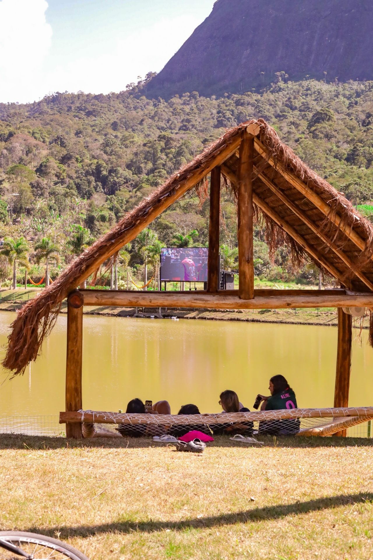 Pessoas descansando sob um abrigo de palha à beira de um lago, com montanhas ao fundo.