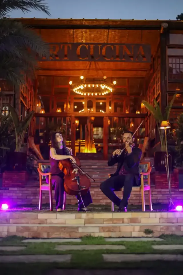 Dois músicos tocando violoncelo e violino no pátio iluminado de um restaurante à noite.