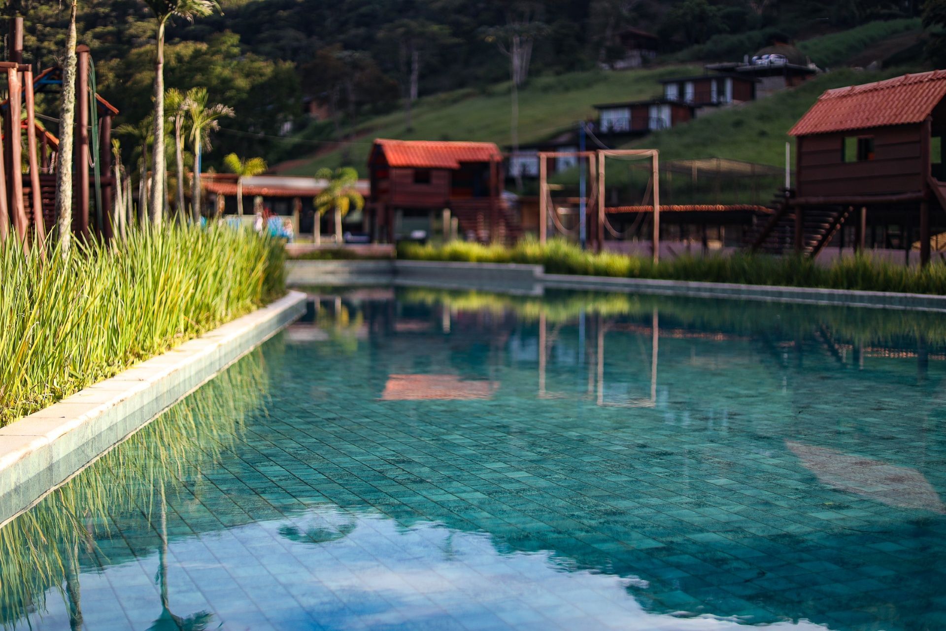 Piscina do resort ao lado de cabanas de madeira e vegetação exuberante, ao pé de uma encosta arborizada.