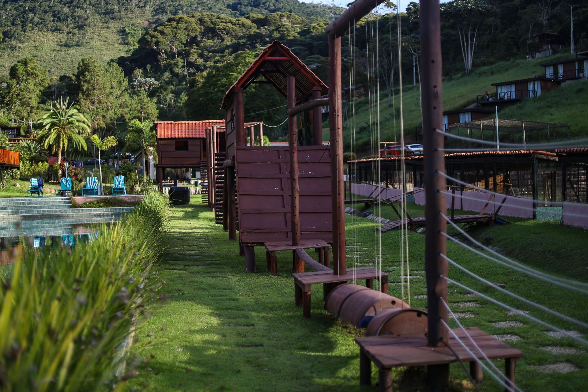 Ponte de corda à beira do lago e passarela de madeira em um parque verde com colinas ao fundo.