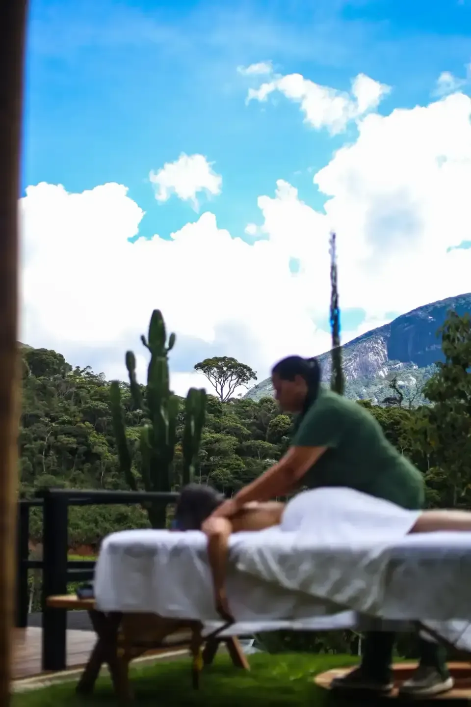 Pessoa massageando alguém em uma mesa ao ar livre, com montanhas e céu azul ao fundo.