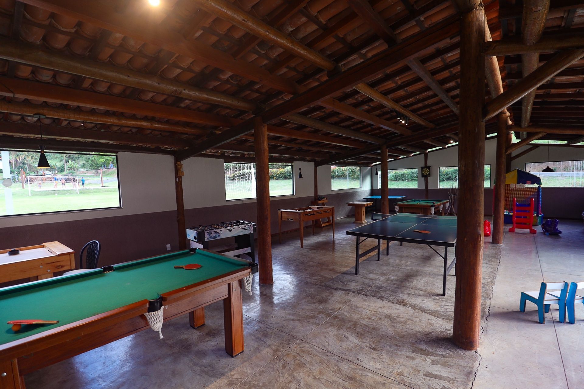 Sala de jogos ao ar livre com mesa de bilhar, mesa de pingue-pongue, TV e vigas de madeira sob um telhado de telhas.