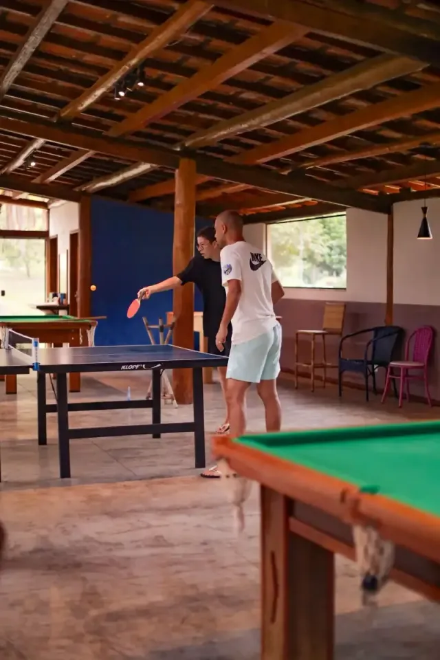 Duas pessoas jogando bilhar em uma sala rústica com uma mesa de bilhar verde e vigas de madeira.
