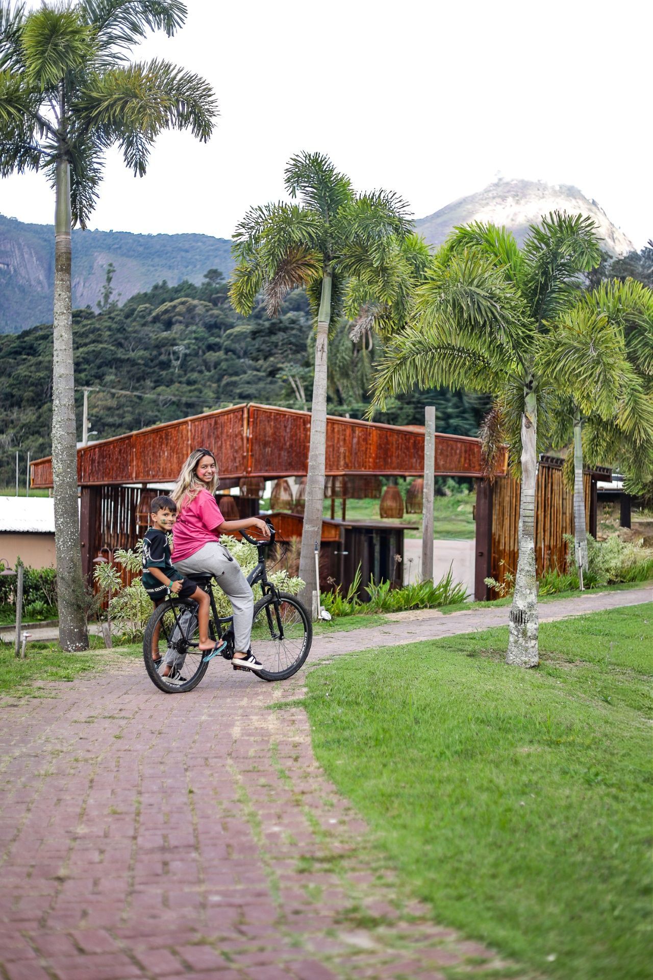 Ciclista de jaqueta rosa pedalando em uma trilha de tijolos ao lado de um parque gramado com palmeiras e montanhas ao fundo.