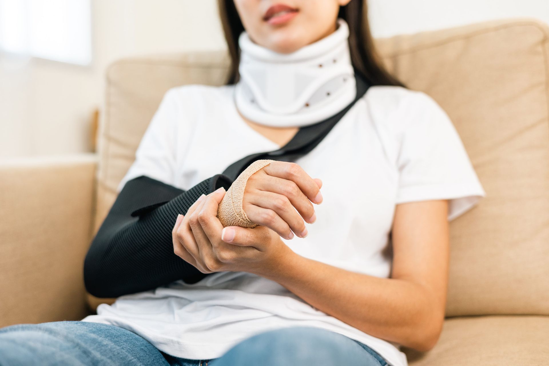 Person wearing a neck brace and arm sling sitting on a couch, holding their wrist.