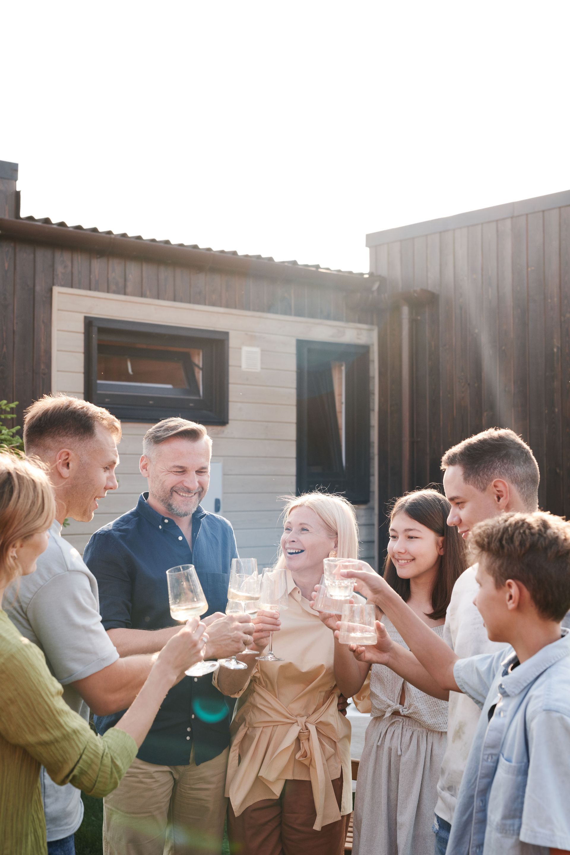 Friends clinking glasses and smiling outdoors beside a wooden cabin in bright sunlight