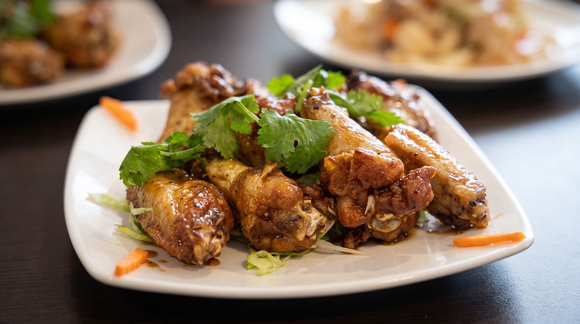 Plate of glazed chicken wings topped with cilantro on a white dish, with more plates blurred in the background