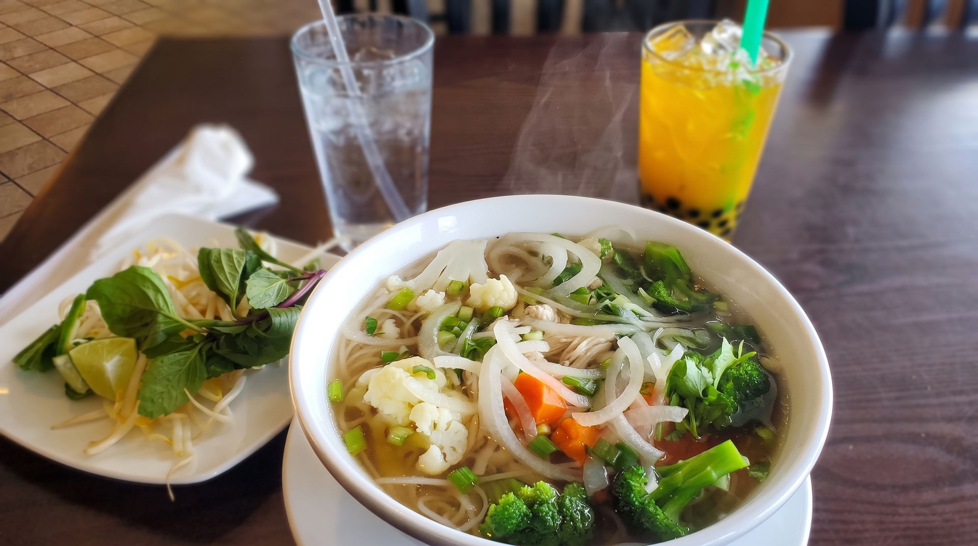 Pho bowl with herbs and vegetables beside a small appetizer plate and two drinks on a wooden table