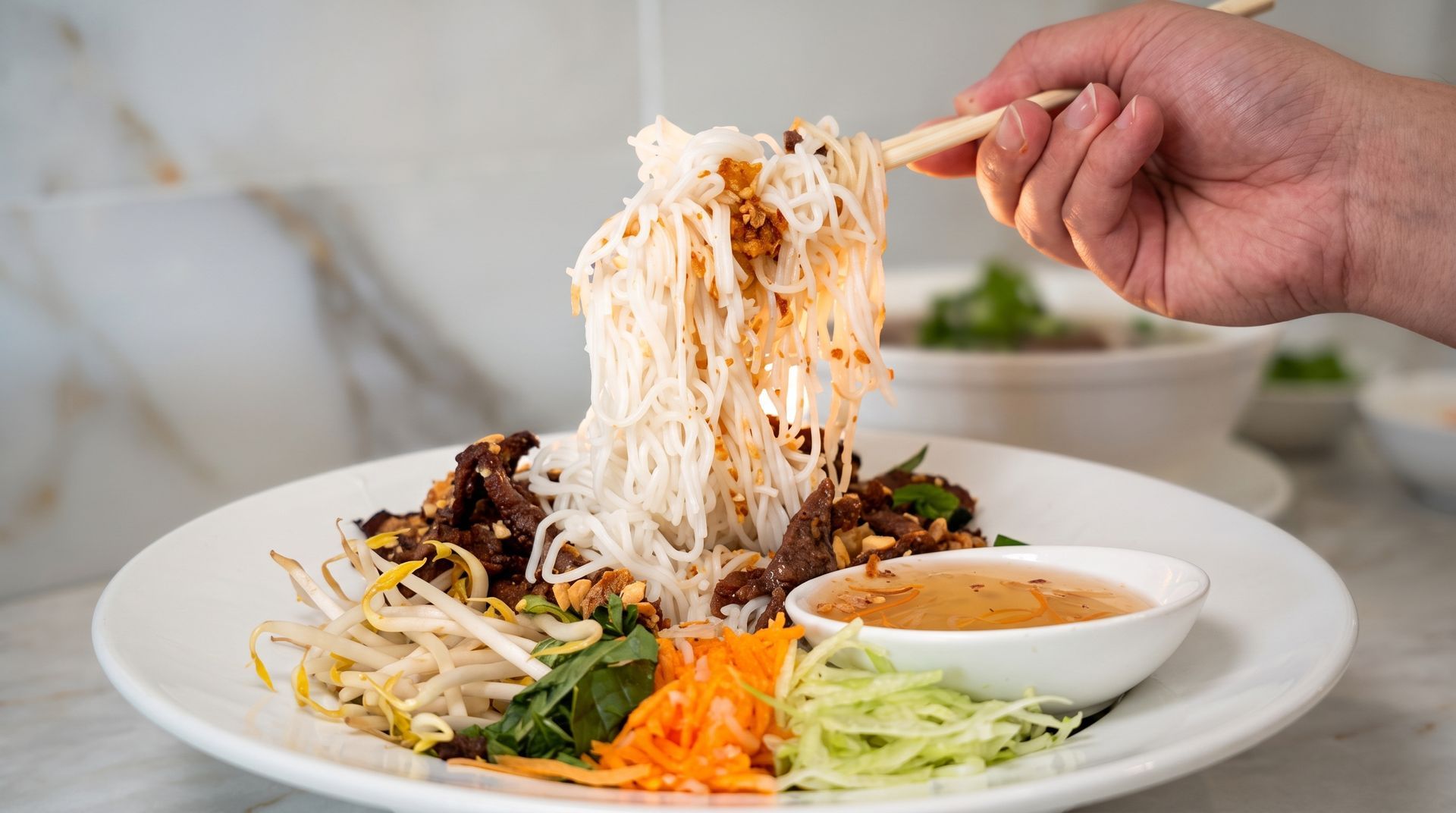 Hand lifting noodles from a plate of Vietnamese noodle salad with herbs and dipping sauce
