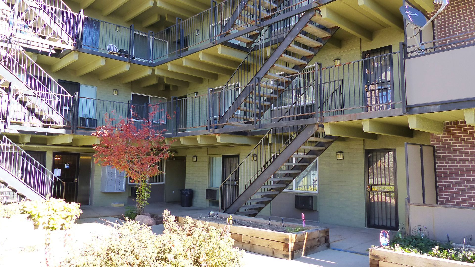 Exterior staircases and balconies of a yellow apartment building, with a small tree and courtyard below