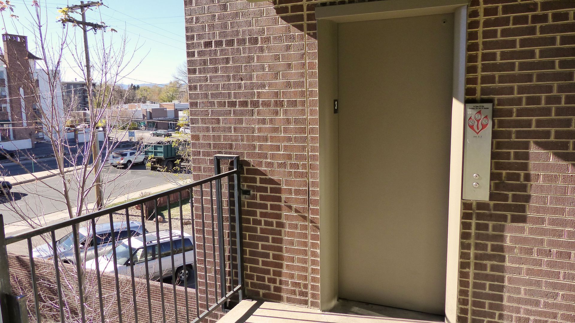Brick apartment entry with beige door and black railing beside a sunny sidewalk
