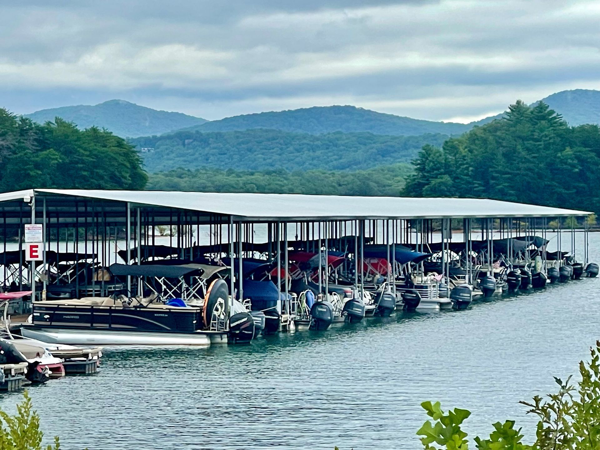 A long dock with covered boat slips on a lake, with mountains in the background under a cloudy sky.