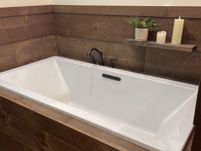 A white rectangular bathtub set in a bathroom with dark brown wood paneling. A small shelf holds candles and a plant.