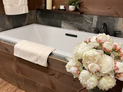 A bathtub with a wooden front, surrounded by dark stone walls, with a bouquet of white flowers and a towel.