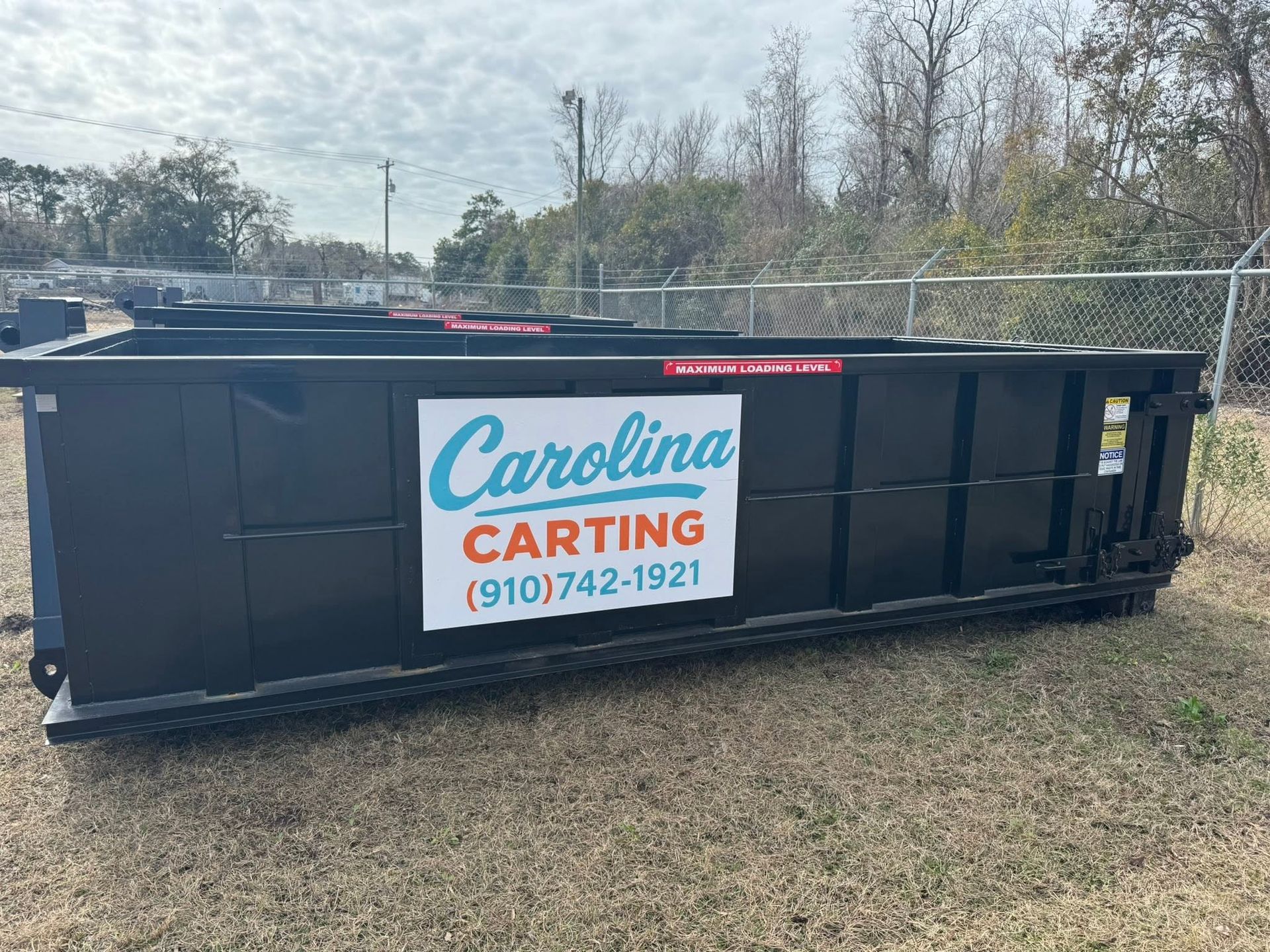 Large black dumpster with Carolina Carting sign in a grassy lot under overcast skies