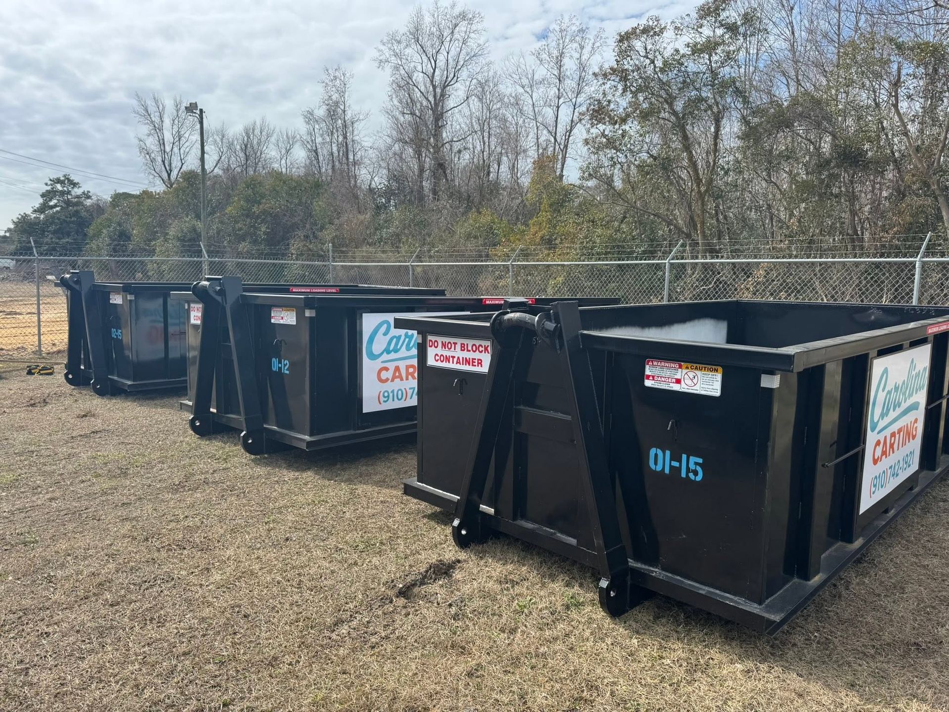 Row of black dumpsters on gravel beside a wooded area under a cloudy sky