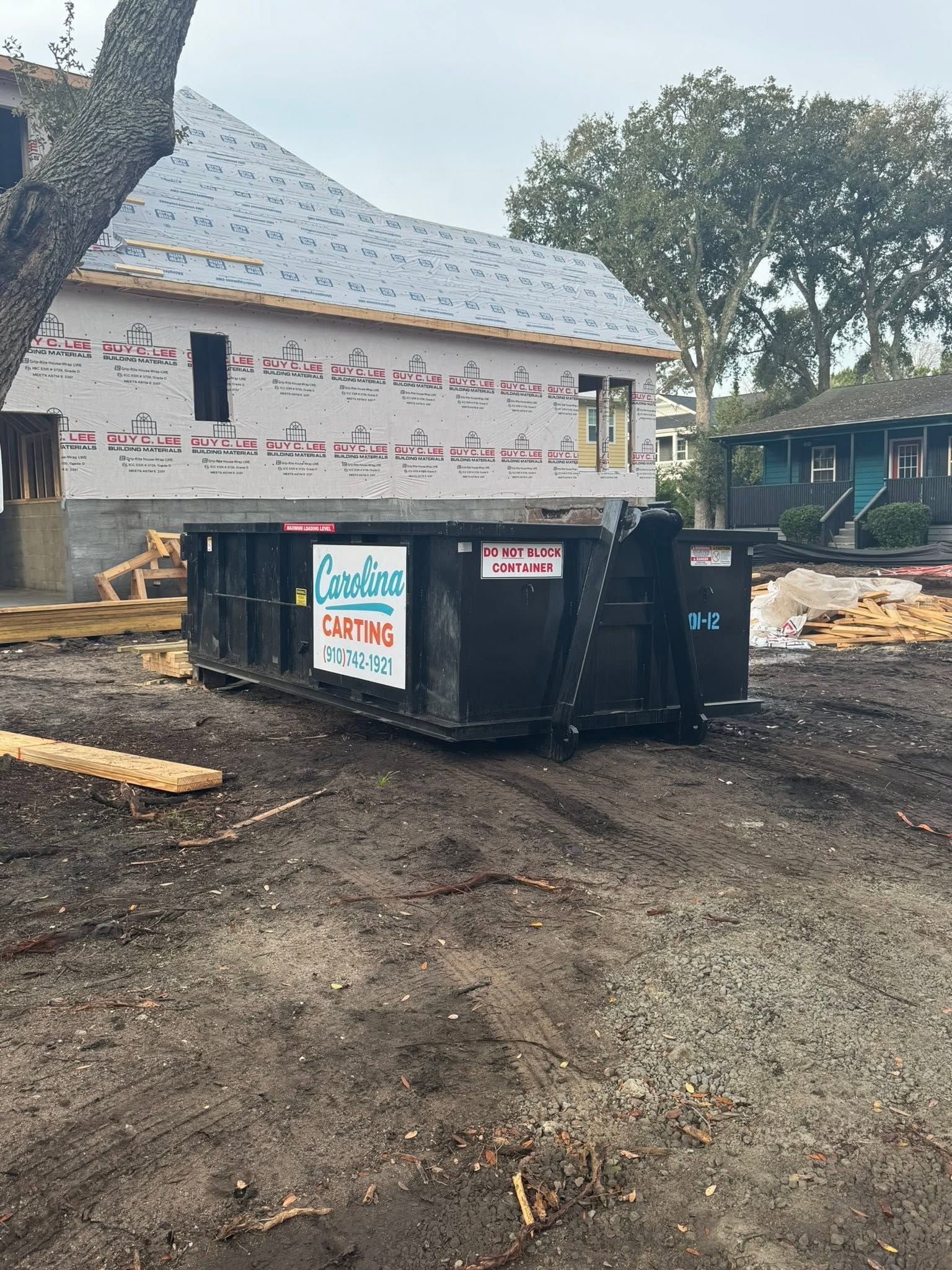 Black dumpster in front of a white building with Arabic writing, on a dirt lot with trees nearby.