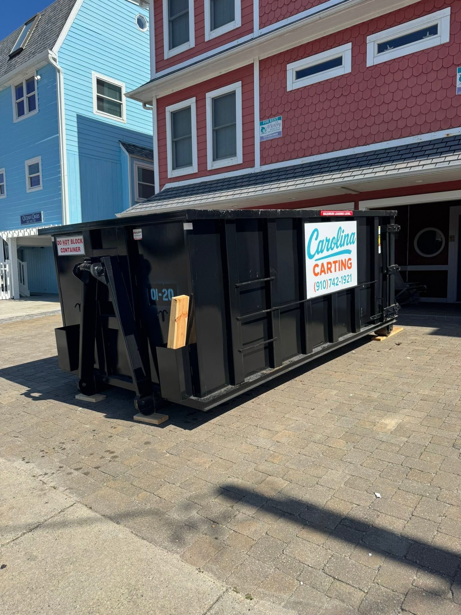 Black dumpster with a sign outside a colorful red and blue building on a sunny street