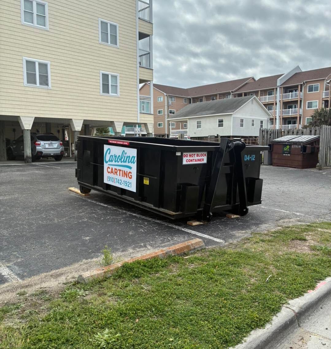 Black dumpster in a parking lot beside apartment buildings and a grassy curb.