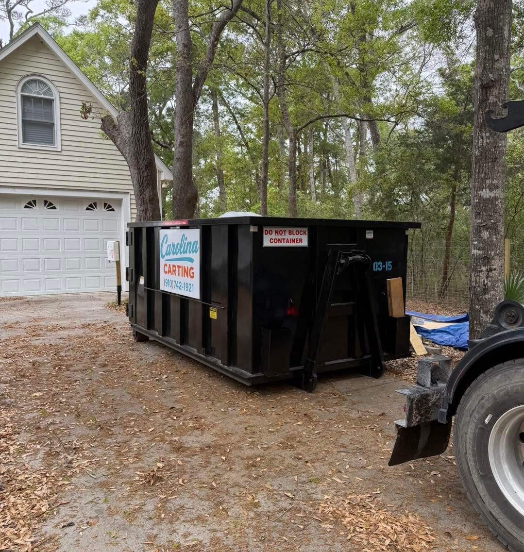 Black dumpster in a wooded driveway beside a garage, with a tractor at right.