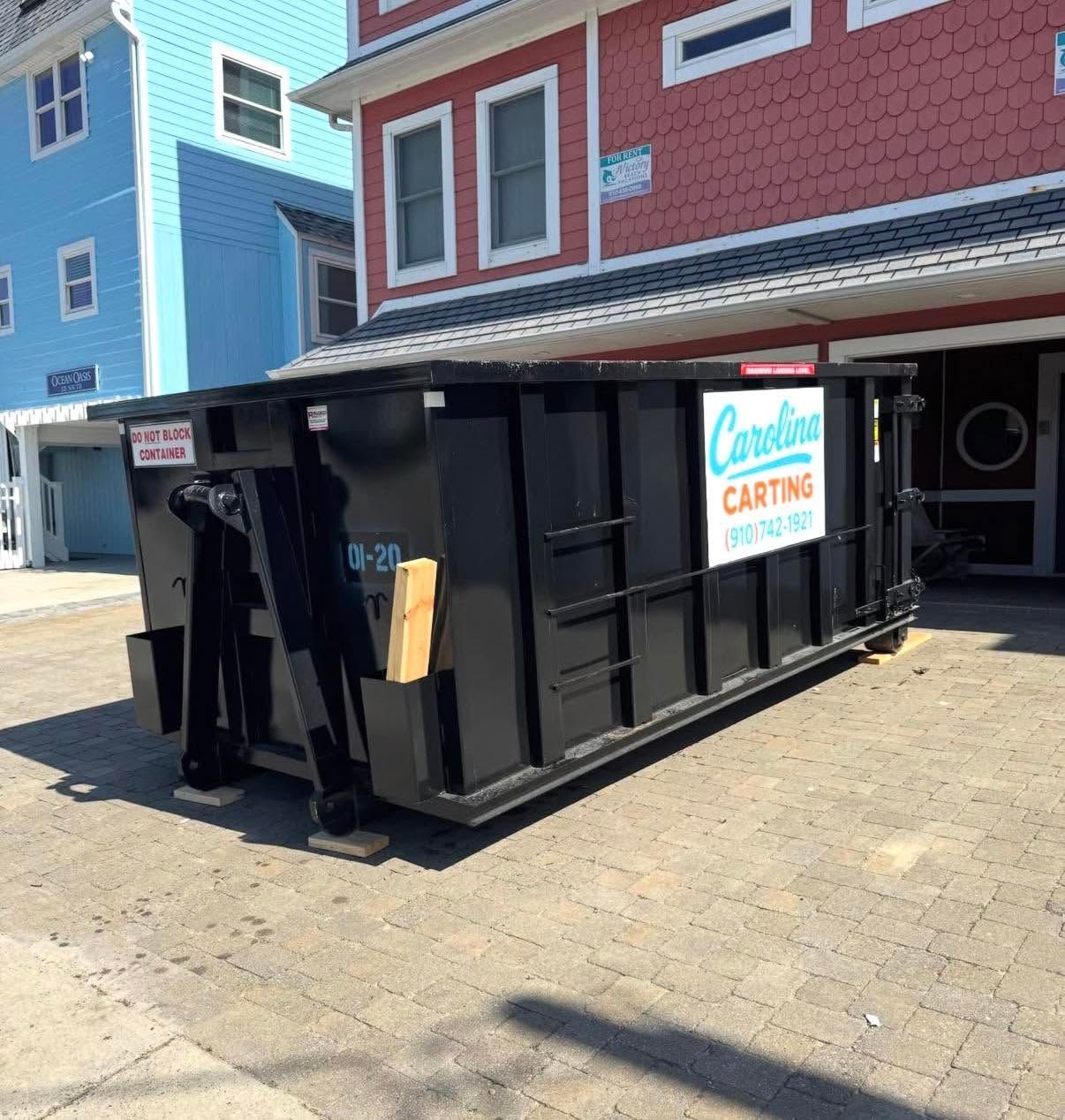 Black dumpster beside a red and blue building with a catering sign on a sunny street