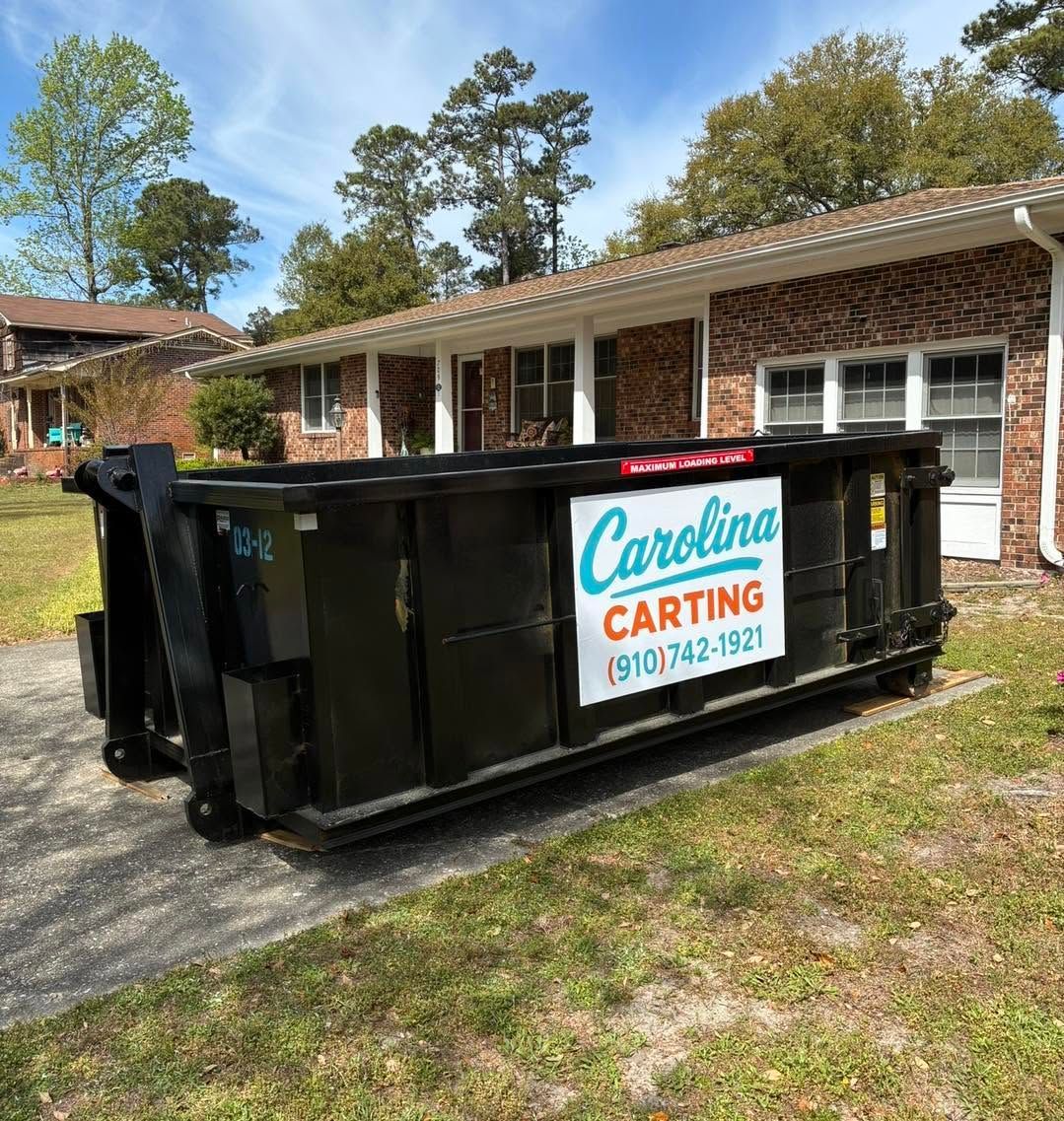 Black roll-off dumpster with Carolina Carting sign in front of a brick house and trees